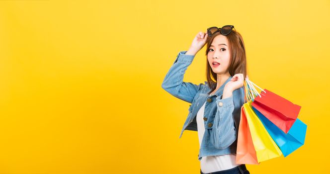 Asian happy portrait beautiful cute young woman teen smiling standing with sunglasses excited holding shopping bags multi color looking camera isolated, studio shot yellow background with copy space