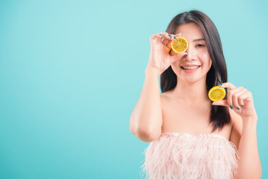 Asian happy portrait beautiful young woman standing smile holding a piece of orange in front of her eye on blue background with copy space for text, healthy lifestyle concept