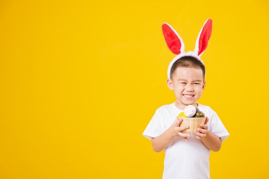 Portrait happy Asian cute little children boy smile standing so happy wearing white T-shirt and bunny ears in Easter festival day holding easter eggs, studio shot on yellow background with copy space