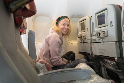 Relaxed cheerful sporty casual young blonde caucasian woman smiling, looking in camera, enjoing extra space while traveling economy class by commercial airplane. Transportation by planes.