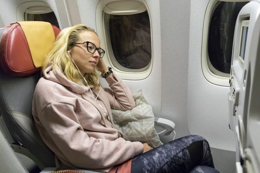 Tired blonde casual caucasian lady napping on uncomfortable seat while traveling by airplane. Commercial transportation by planes.