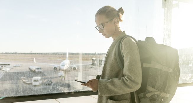 Young woman standing near airport gates window holding cellphone in her hands, wearing travel backpack and walking to lounge area. Female traveler search online map at the airport.