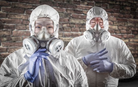 Chinese Woman and Man In Gas Masks, Goggles and Hazmat Suites Against Brick Background.