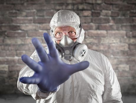 Man Wearing Hazmat Suit, Protective Gas Mask and Goggles Reaching Out With Hand Brick Wall Behind.