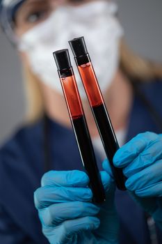 Female Lab Worker Wearing Medical Face Mask Holds Test Tubes of Blood Against Dark Background.