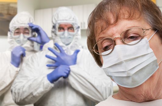 Senior Adult Woman Wearing Medical Face Mask In Doctor Office With Nurses In HAZMAT Suits Behind.