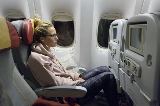 Sporty casual young blonde caucasian lady watching movie while traveling by airplane by the window. Commercial transportation by planes.