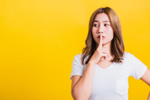 Asian Thai happy portrait beautiful cute young woman standing wear white t-shirt making finger on lips silent quiet gesture looking to side, studio shot isolated on yellow background with copy space