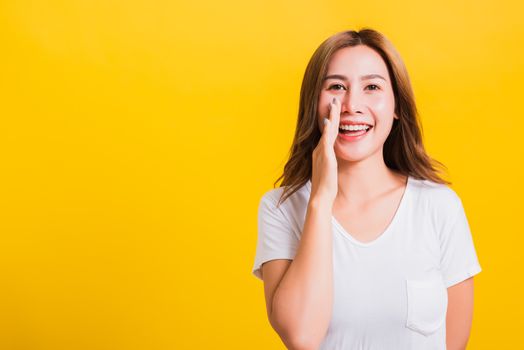 Asian Thai happy portrait beautiful young woman standing wear t-shirt hold a hand on mouth talking whispering secret rumor looking to camera isolated, studio shot on yellow background with copy space