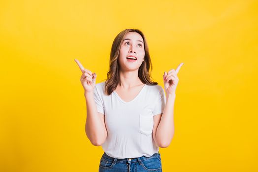 Asian Thai happy portrait beautiful cute young woman standing wear white t-shirt makes gesture two fingers point upwards above looking above, studio shot isolated on yellow background with copy space