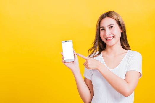 Asian Thai portrait happy beautiful young woman smile standing wear t-shirt making finger pointing on smartphone blank screen looking to camera isolated, studio shot yellow background with copy space