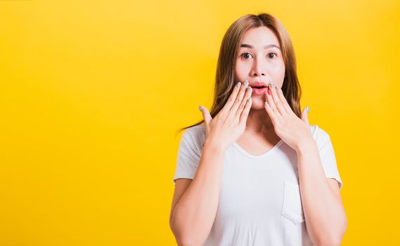 Asian Thai happy portrait beautiful cute young woman standing amazed, shocked afraid wide open mouth gesturing hand palms looking to camera, studio shot isolated on yellow background with copy space