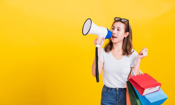 Portrait Asian Thai beautiful happy young woman smiling hold shopping bags multi-color and shouting in megaphone her looking to camera, studio shot isolated on yellow background, with copy space