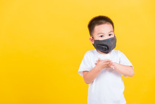 Asian Thai happy portrait cute little cheerful child boy wearing mask protective for covid-19 or pm2.5 dust he clean hand with sanitizing gel, studio shot isolated on yellow background with copy space