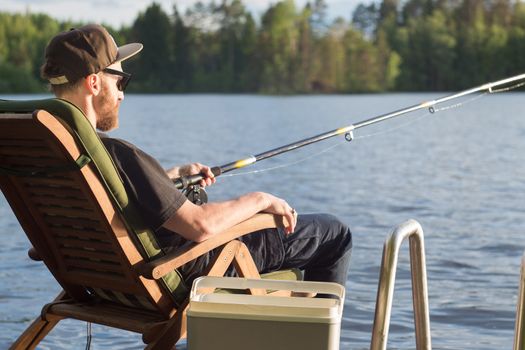 Mature man fishing from wooden pier near cottage on lake in Finland at summer