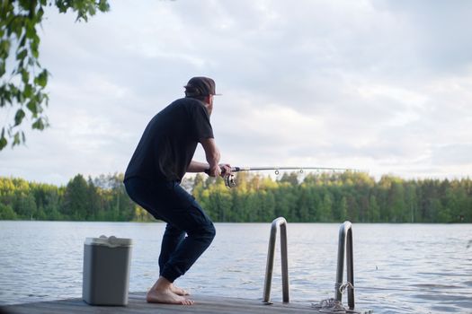 Mature man fishing from wooden pier near cottage on lake in Finland at summer