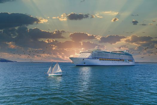 Huge, white, luxury cruise ship anchored in blue water with sailboat off starboard bow