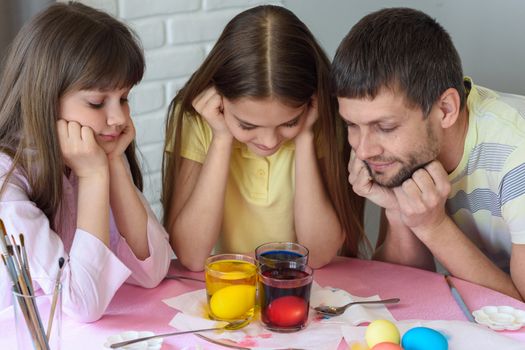 Dad and two daughters are watching how the eggs are painted in glasses with dyes