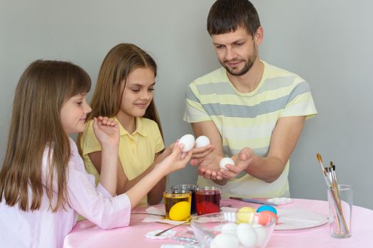 Dad and daughters sit at the table and paint eggs for Easter