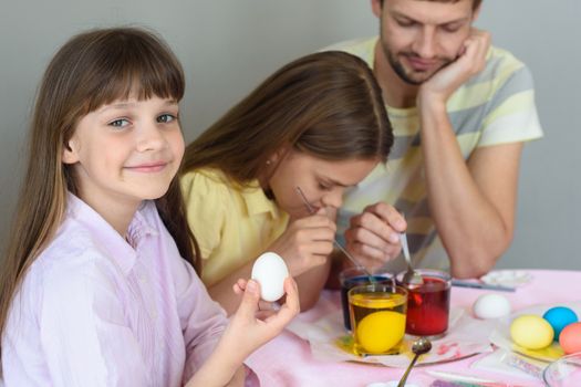 A girl with an egg in her hands looked into the frame, in the background the family paints eggs in glasses with dyes