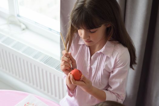 Girl paints an easter egg sitting by the window in a home environment