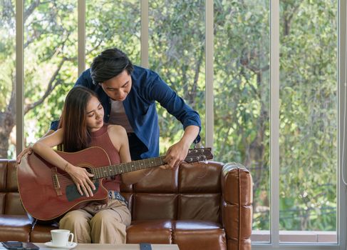 Young couples spend holidays in the living room. The young man wears comfortable clothes, teach his girlfriend to play a guitar. Asian women focus on music lesson.