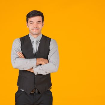 A young businessman in a gray shirt and a black vest fold his arms over the chest with confidence. The concept of investing in a successful business. Portrait on a yellow background with studio lights