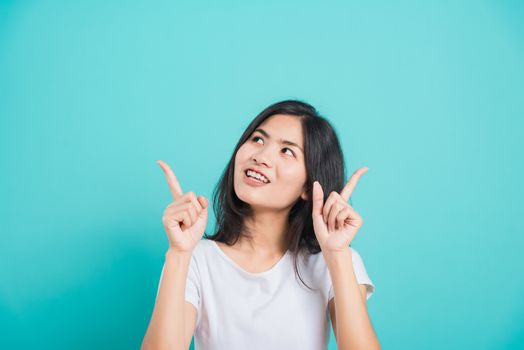Portrait Asian beautiful young woman wearing white T-shirt standing smile white teeth, She pointing finger up and looking up, shoot photo in studio on blue background, There was copy space.