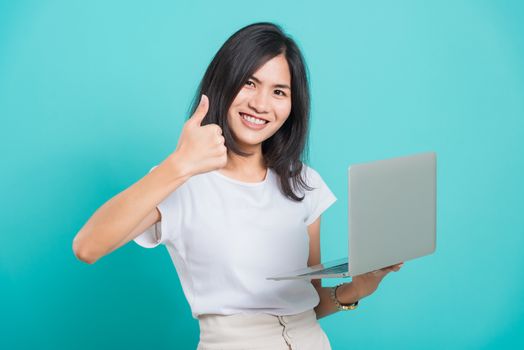 Asian beautiful young woman smile white teeth standing to holding laptop computer and showing thumb up while shoot photo in studio on blue background with copy space for text