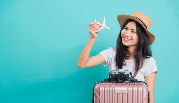 Traveler tourist happy Asian beautiful young woman, holidays travel concept, her holding fly toy aircraft her have suitcase bag and photo mirrorless camera, shoot photo in studio on blue background