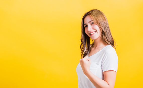 Asian Thai happy portrait beautiful cute young woman stand wear t-shirt makes raised fists up celebrating her winning success looking to camera, studio shot isolated yellow background with copy space