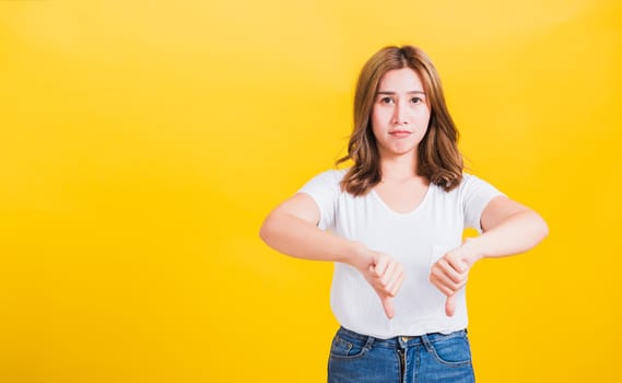 Portrait Asian Thai beautiful young woman unhappy, a negative gesture showing finger thumbs down or dislike sign, studio shot isolated on yellow background, There was copy space, rejection concept