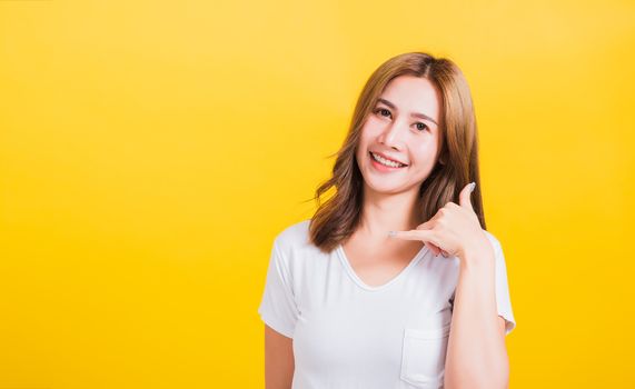 Portrait Asian Thai beautiful happy young woman wear white t-shirt standing smiling white teeth, making call me gesture by finger hand isolated on yellow background, with copy space