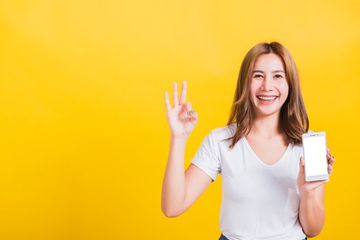 Portrait Thai Asian beautiful happy young woman stand smile, holding mobile phone blank screen and showing OK gesture, shoot photo in studio focus face isolated on yellow background, with copy space