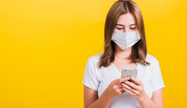 Portrait Asian Thai beautiful happy young woman wearing mask protection, reading news update on smartphone health care, studio shot isolated on yellow background, with copy space