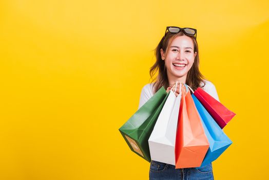 Asian Thai portrait happy beautiful cute young woman smiling stand with sunglasses excited holding shopping bags multi color looking camera, studio shot isolated yellow background with copy space