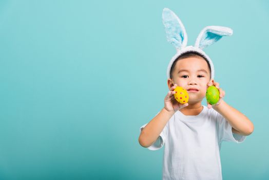 Asian cute little child boy smile beaming wearing bunny ears and a white T-shirt, standing using hand holding and show colored easter eggs on blue background with copy space