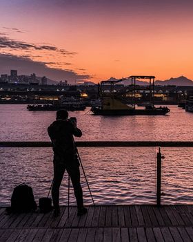 The silhouette of people, tripod, glass fence near Hong Kong river in downtown district at sunset