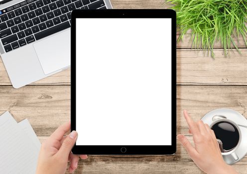 Laptop computer, green plants, coffee on the office wooden desk and human hand holding big tablet