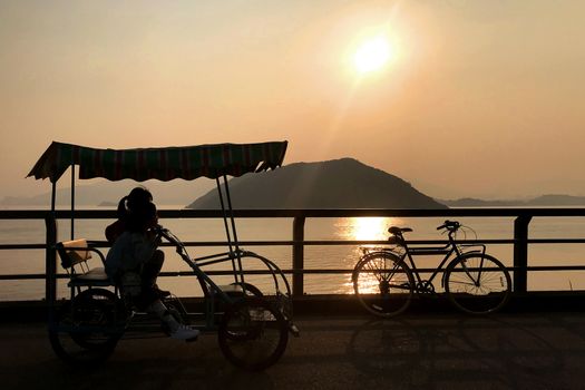 The silhouette of retro bicycle, family bike, fence, ocean, mountain and sun at sunset