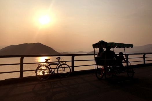 The silhouette of retro bicycle, family bike, fence, ocean, mountain and sun at sunset