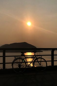 The silhouette of retro bicycle, fence, ocean, mountain and sun at sunset