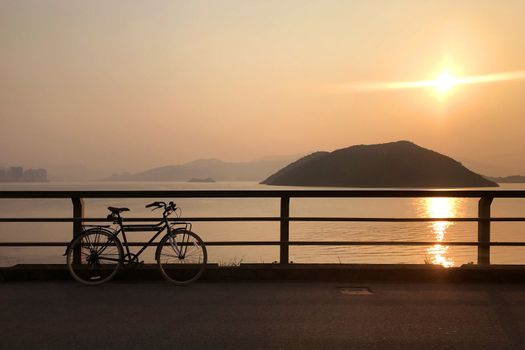 The silhouette of retro bicycle, fence, ocean, mountain and sun at sunset