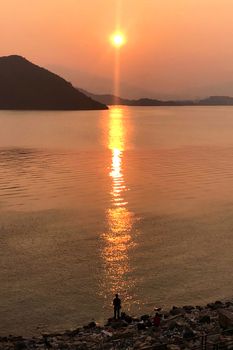 The vertical silhouette of fishing man, ocean, mountain and sun at sunset