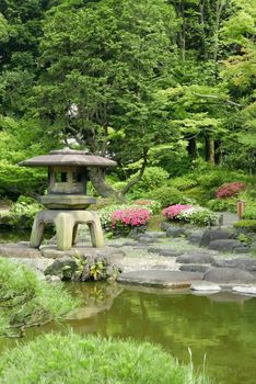Vertical Japanese outdoor stone lantern, flower plants and lake in the zen garden