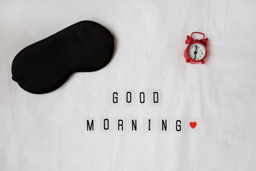 Silk black sleep mask, inscription Good morning, red clock on white rumpled sheets. Top view, flat lay. Horizontal. Concept of rest, awakening, sleep. For social media, blog. Minimal style.