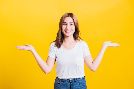 Asian Thai happy portrait beautiful cute young woman standing wear t-shirt holding something on palm two hands away side looking to camera, studio shot isolated on yellow background with copy space