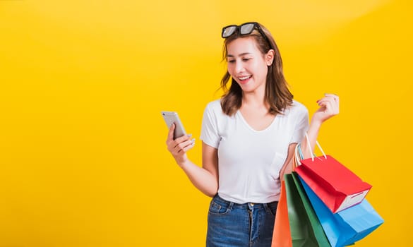 Portrait happy Asian Thai beautiful young woman smile white teeth stand wear t-shirt, She holding shopping bags and using app in mobile phone, studio shot isolated on yellow background with copy space