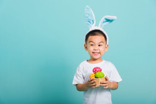 Asian cute little child boy smile beaming wearing bunny ears and a white T-shirt, standing to hold a basket with full Easter eggs on blue background with copy space