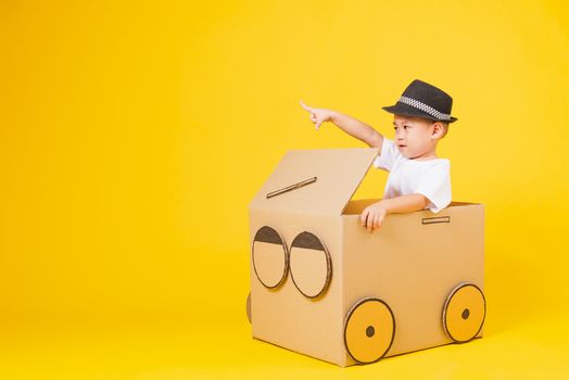 Portrait happy Asian cute little children boy smile so happy wearing white T-shirt driving car creative by cardboard and pointing finger, studio shot on yellow background with copy space